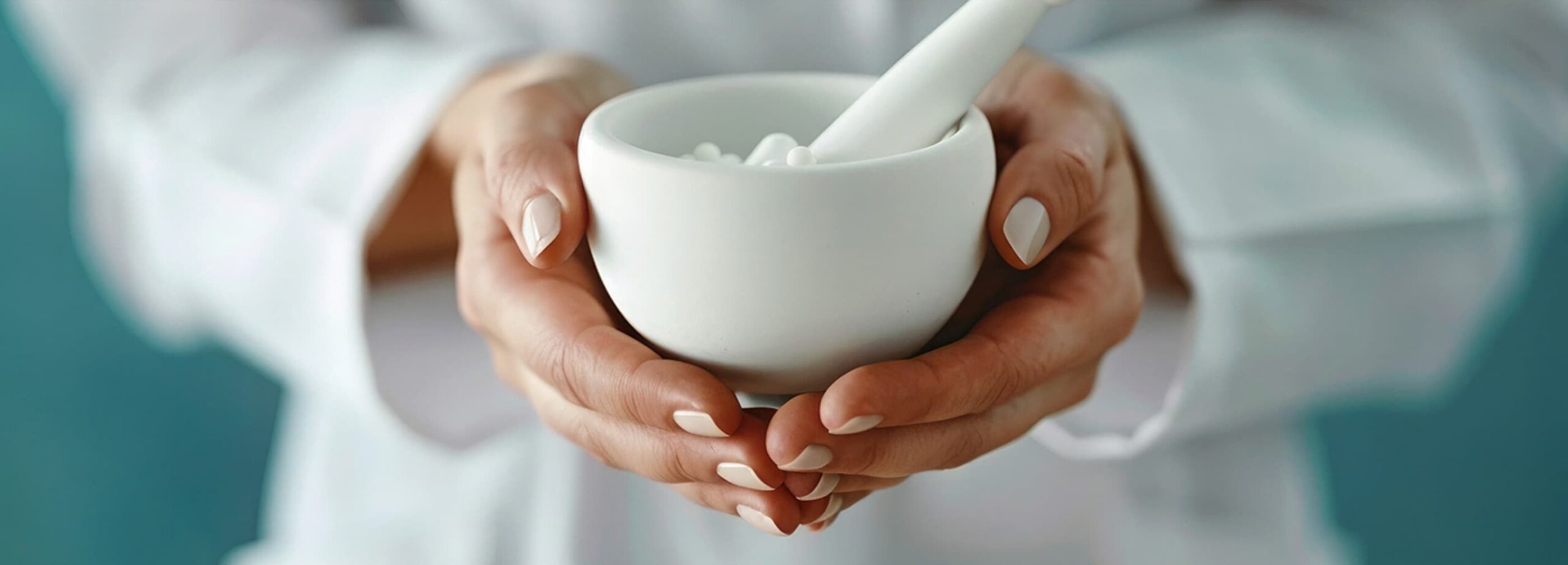 A close-up of a person in a white lab coat holding a white ceramic mortar and pestle with small white pills inside. The hands are gently cradling the mortar, and the background is blurred with a soft teal hue.