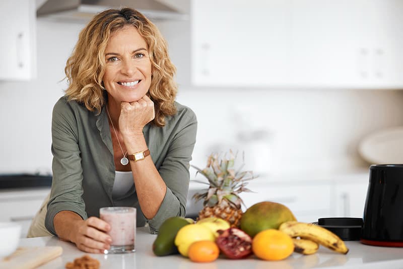 Woman leaning on kitchen counter drinking a smoothie with fruit arranged at her side.