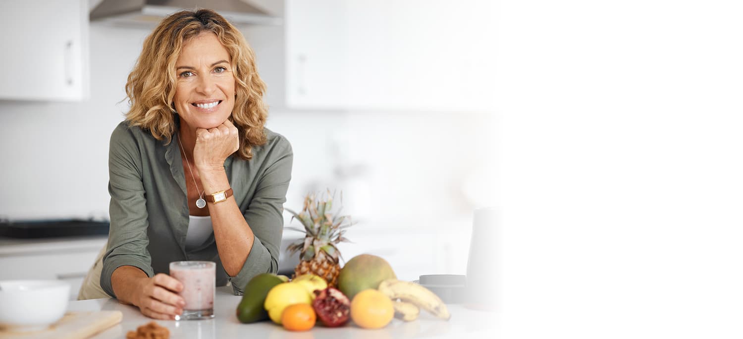 Woman leaning on kitchen counter drinking a smoothie with fruit arranged at her side.